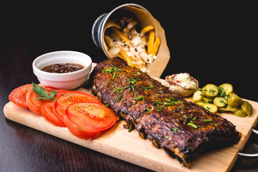 ribs on a wooden board surrounded by tomatoes, barbecue sauce, fries and pickles