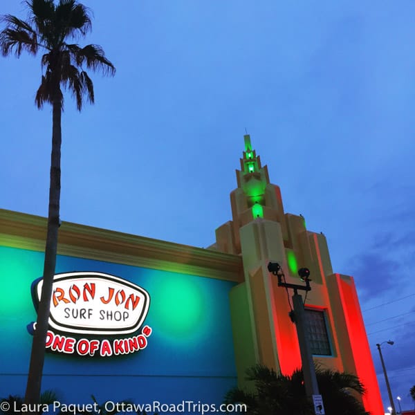 Facade of Ron John Surf Shop illuminated with multi-coloured floodlights lights at dusk.