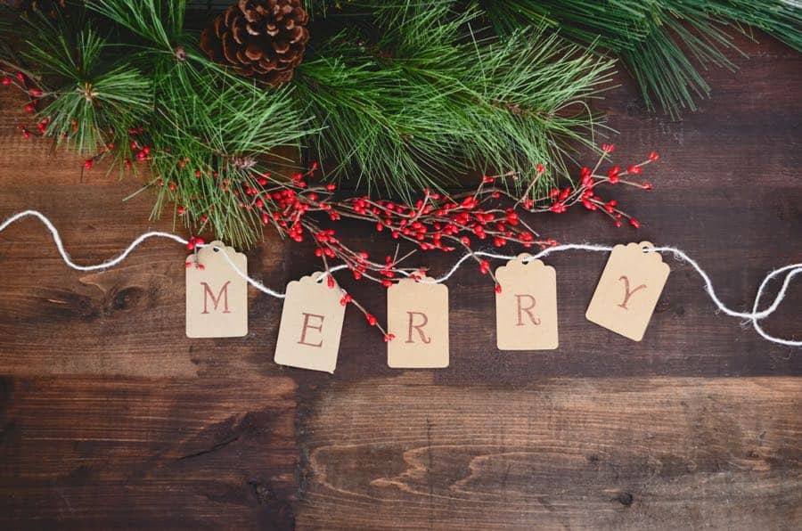 tags spelling out merry on a wooden table with pine boughs and holly berries.