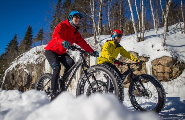 Cyclist in red jacket and cyclist in yellow coat on fat bikes on snowy, hilly trail lined with birch trees in the Mont Tremblant area of Quebec.
