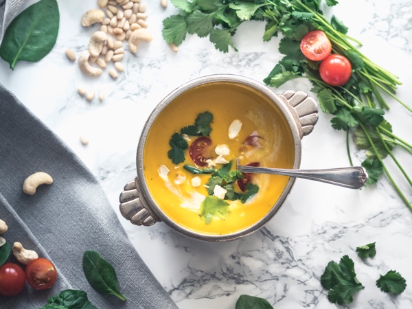 Soup bowl with bright orange soup surrounded by herbs and vegetables. Photo by Nathalie Jolie on Unsplash.