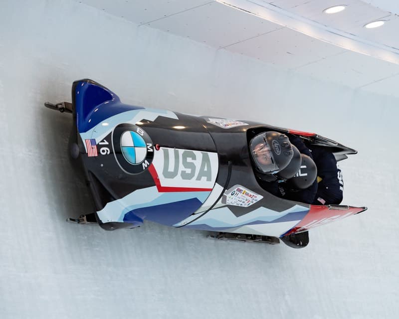 racer in a red, white, blue and black bobsled on an icy track in lake placid, new york.