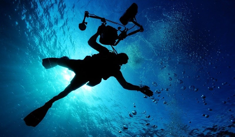 scuba diver with video equipment silhouetted against blue water and sun