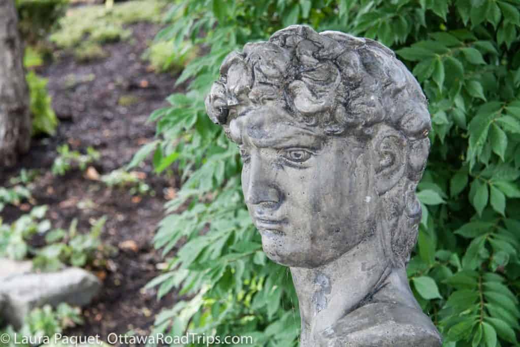 classical style stone bust of a man's head with green plants in background at Shakespeare's Gardens in Prescott, Ontario