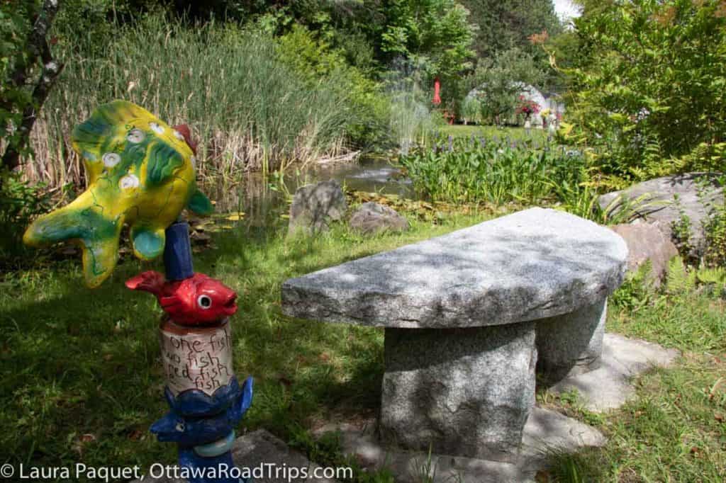 colourful ceramic fish sculpture next to stone bench with pond and fountain in background, at rideau woodland ramble near merrickville.