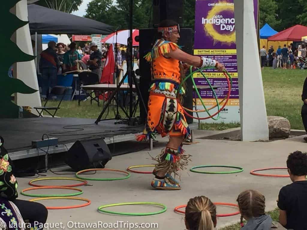 hoop dancer in orange traditional clothing performing on a stage with children sitting watching.