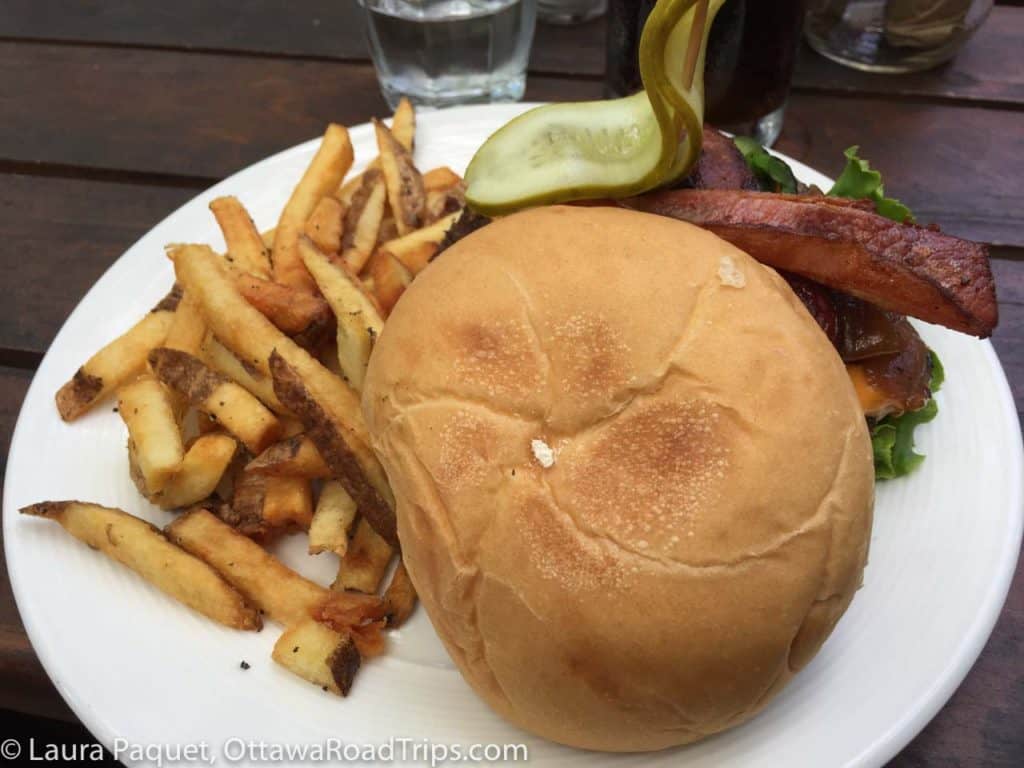 large hamburger with french fries and a pickle on a white plate on the patio at the rocky river cafe in perth, ontario.