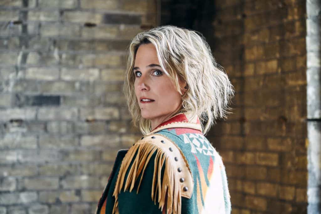 blonde woman in a fringed jacket, looking over her shoulder at the camera