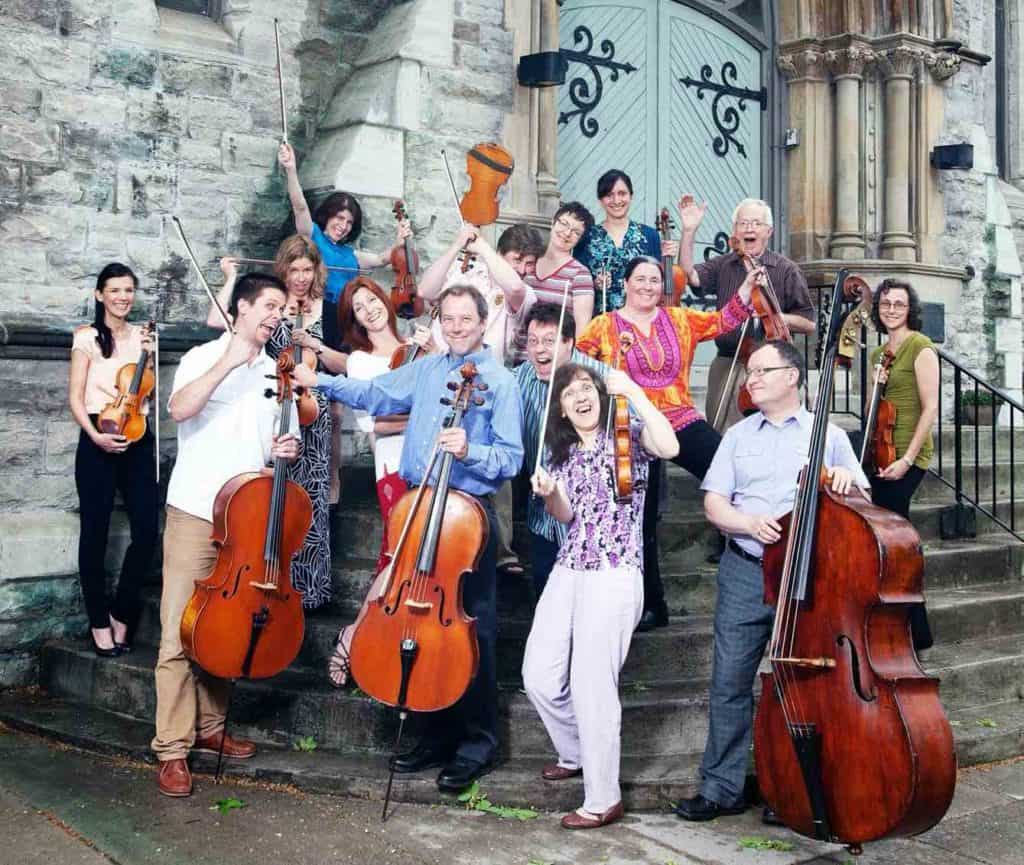 About a dozen musicians, some holding stringed instruments, laughing on the steps of a church