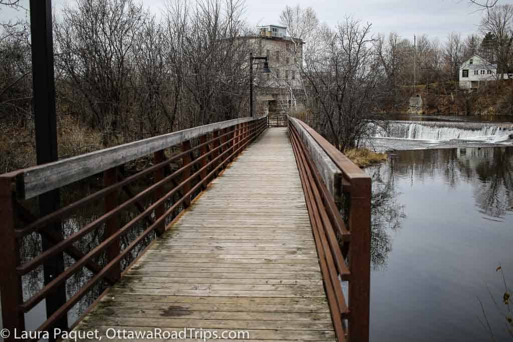 wooden bridge over water with stone mill and waterfall in background in almonte, ontario.