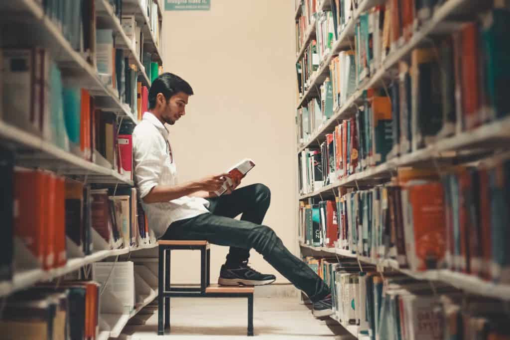 A man sitting on a step stool reading a book in an aisle lined with library bookshelves.