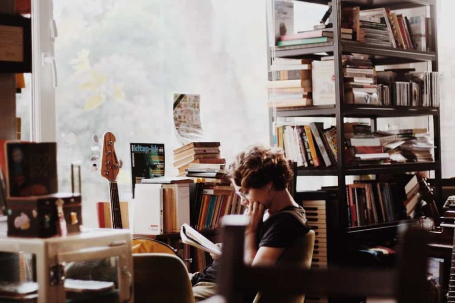 Woman reading a book near a window and packed bookshelf.