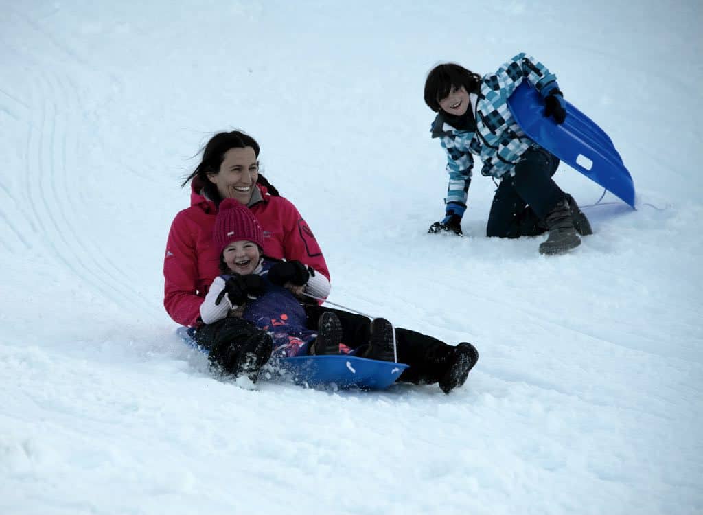 Woman and two kids sledding down a snowy hill.