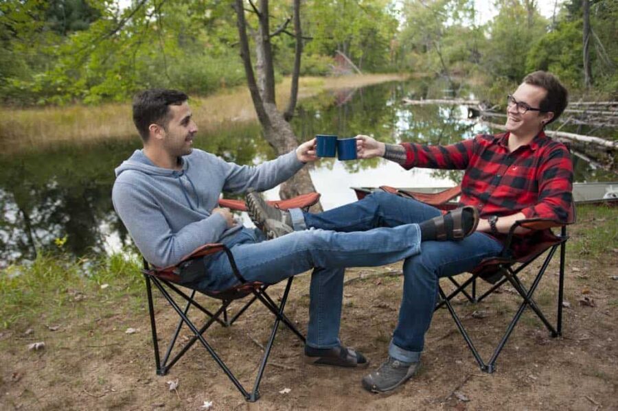 men in camp chairs with coffee mugs next to a lake beach.