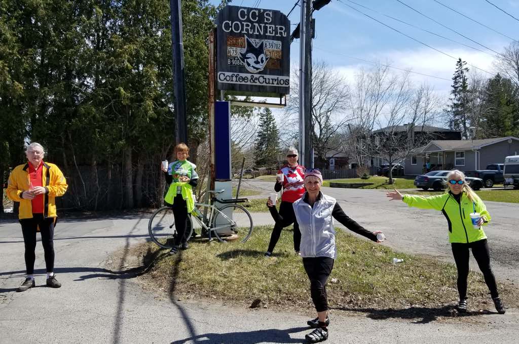 Three cyclists in front of a cafe sign on a rural road, in spring.