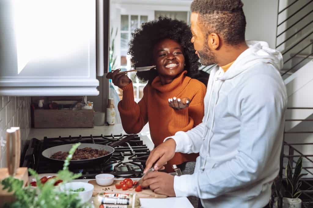 man chopping vegetables as woman tastes something from a spoon in a home kitchen