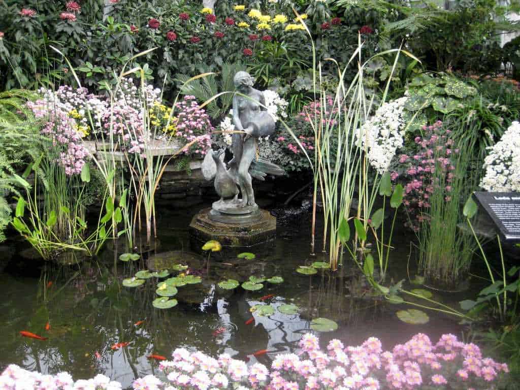 a small sculpture of a woman pouring water in a pond surrounded by flowers