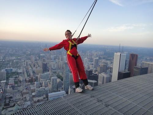 woman in red jumpsuit leaning back from a metal catwalk with toronto skyline below