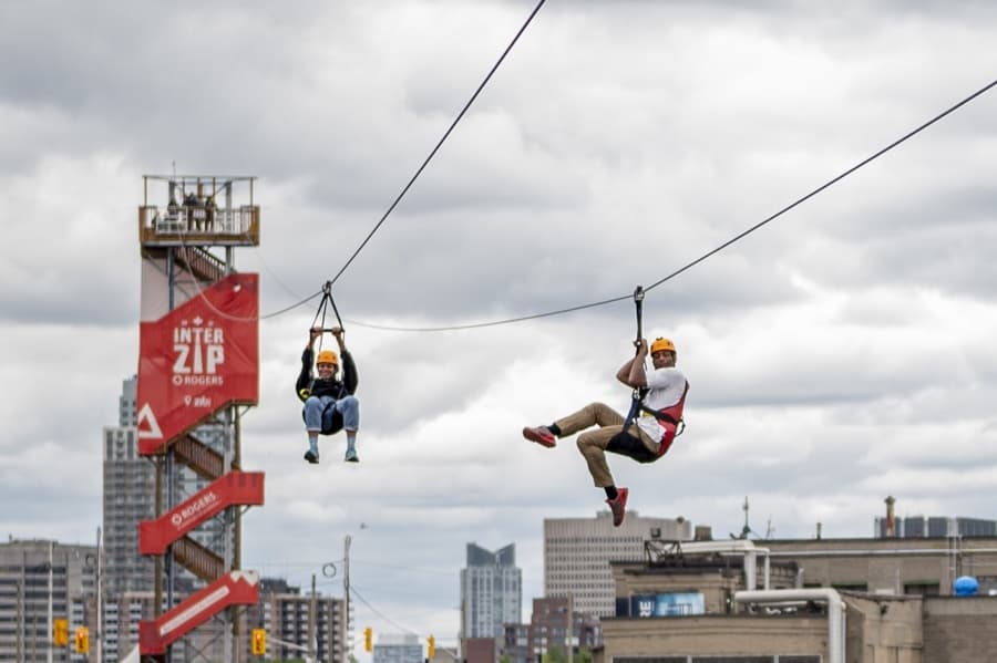 a woman and man hanging from parallel zip lines on interzip rogers in ottawa, with red tower and city buildings in background.