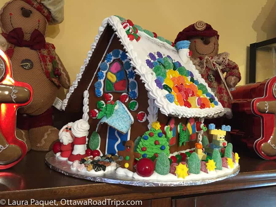 large, heavily decorated gingerbread house on a shelf, surrounded by gingerbread stuffed toys.