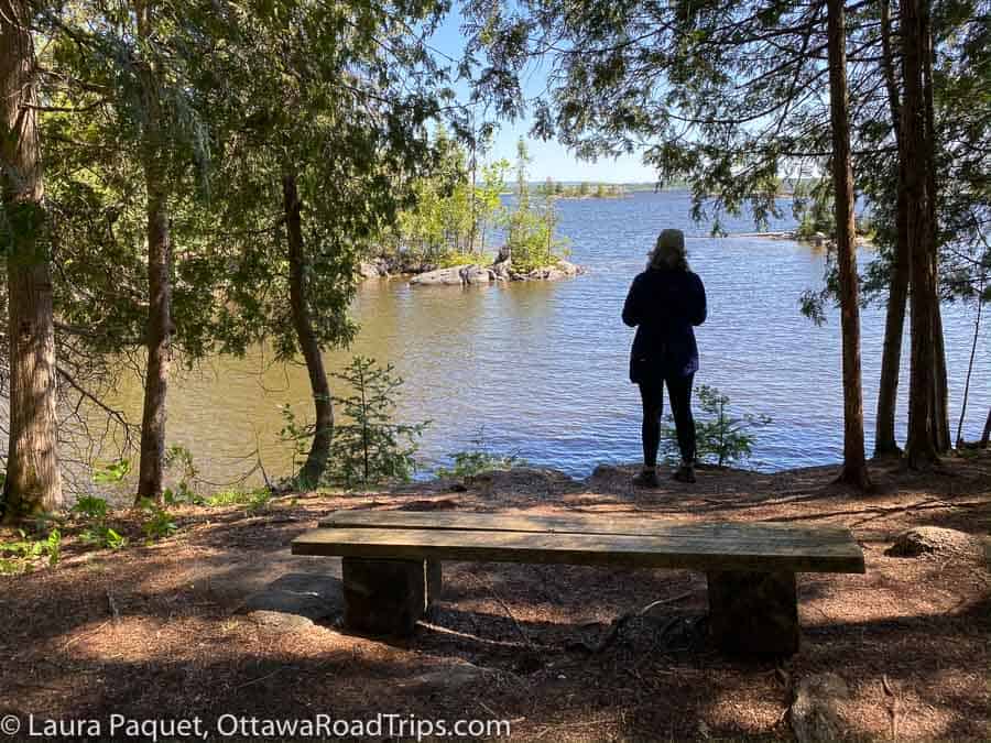 silhouette of a woman looking out over river dotted with rocky island, surrounded by coniferous trees and with a rustic wooden bench behind her.