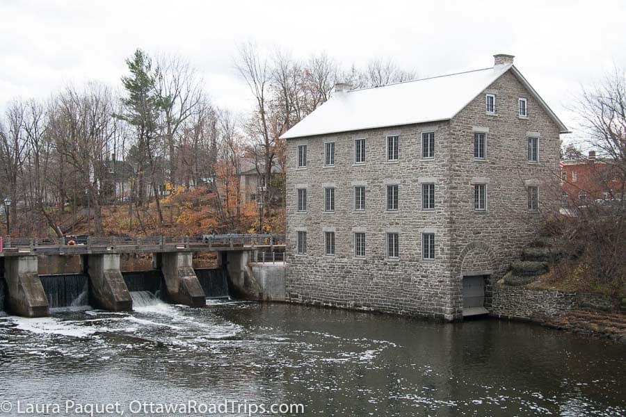 large grey stone mill with small windows next to a dam, surrounded by late fall trees with coloured leaves.