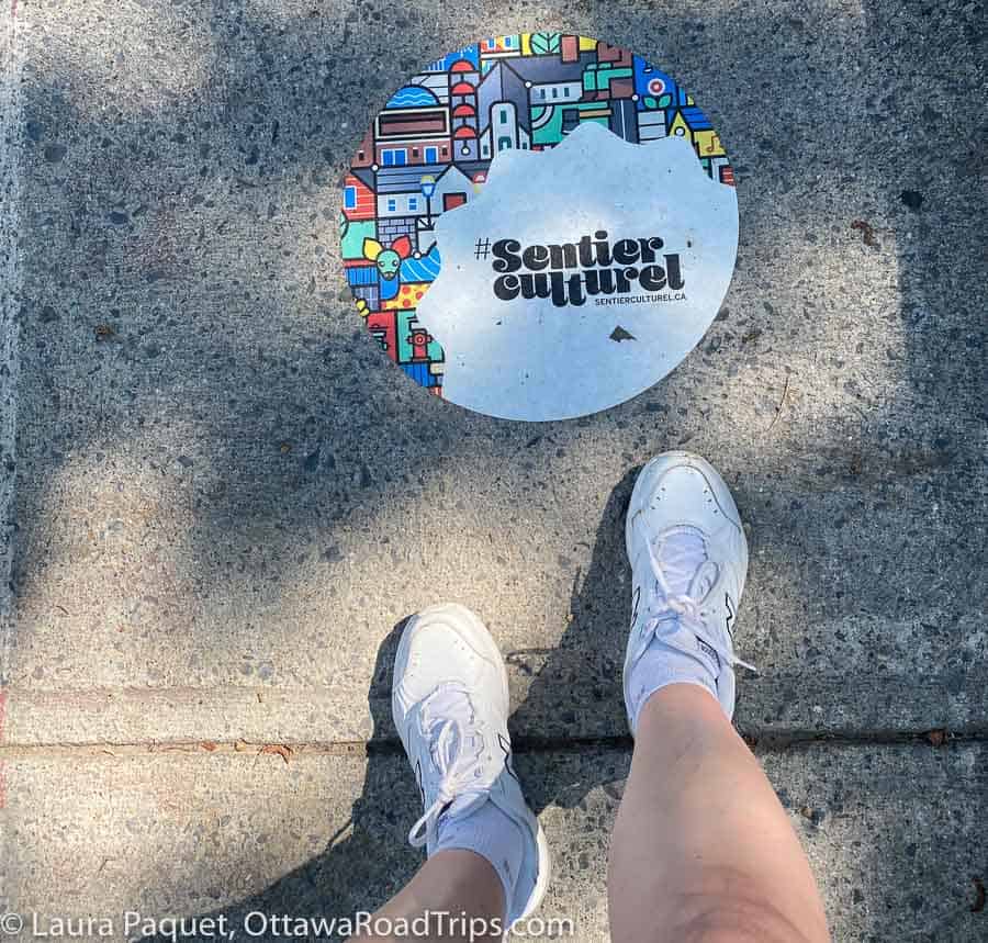 round sentier culturel sign on a grey sidewalk, with a pair of feet in white running shoes.