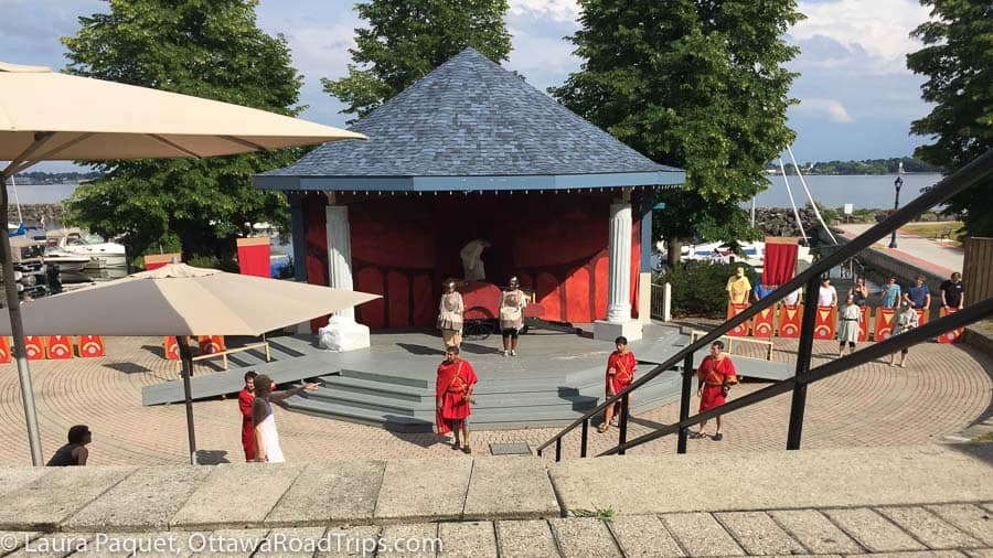 Actors in red and white togas on a semi-circular outdoor stage made of paving stones and wooden risers, with tall green trees and St. Lawrence River in background.