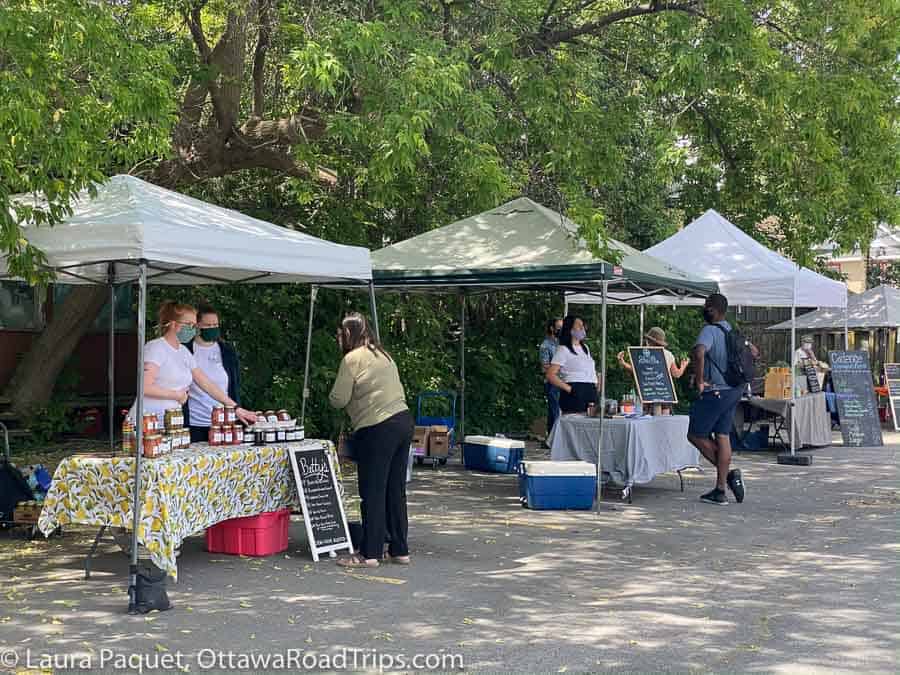 Customers shopping for jarred foods displayed on tables under tents and trees in a parking lot.