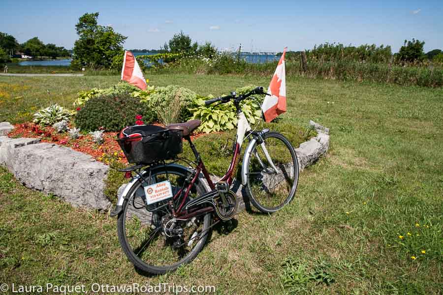 bike parked near a small flower garden with canadian flags and lake ontario in background on wolfe island