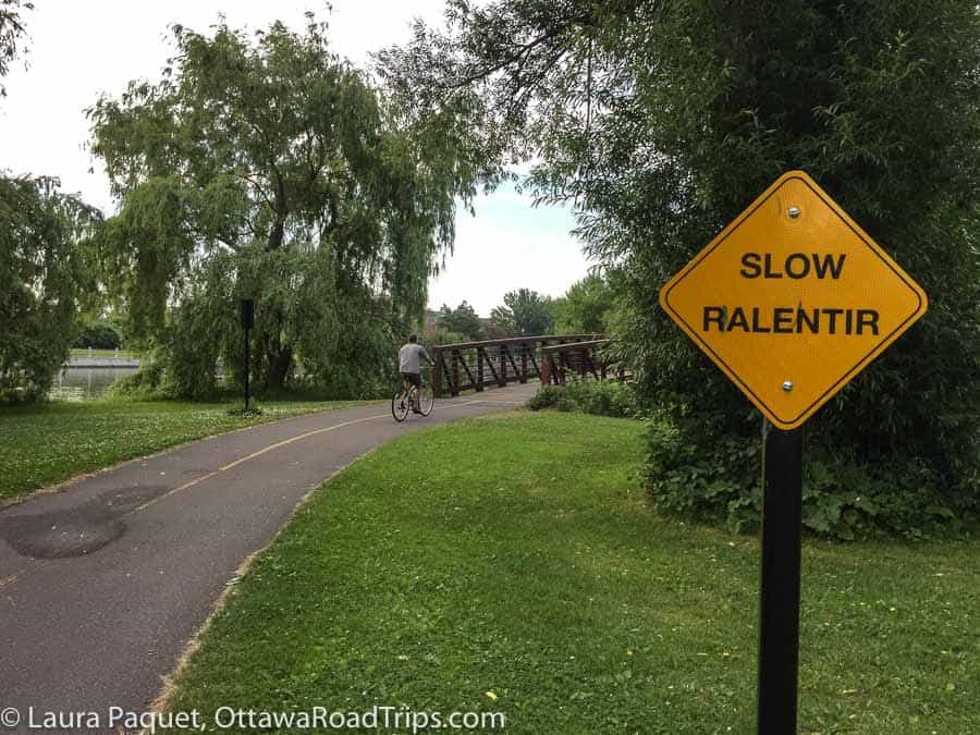 cyclist on a paved path about to cross a wooden bridge, with a sign reading "slow ralentir" in the foreground.