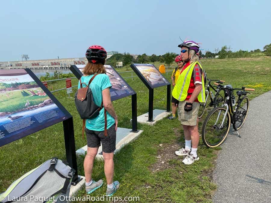 three cyclist looking at large illustrated panels outdoors with grey dam and river in background