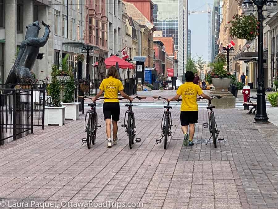 two women in yellow shirts wheeling four bikes along sparks street in ottawa