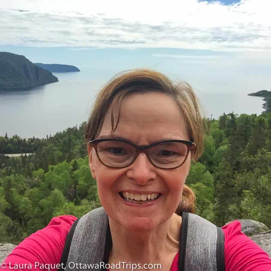 woman in pink shirt smiling at camera, with forest, road and lake superior in background