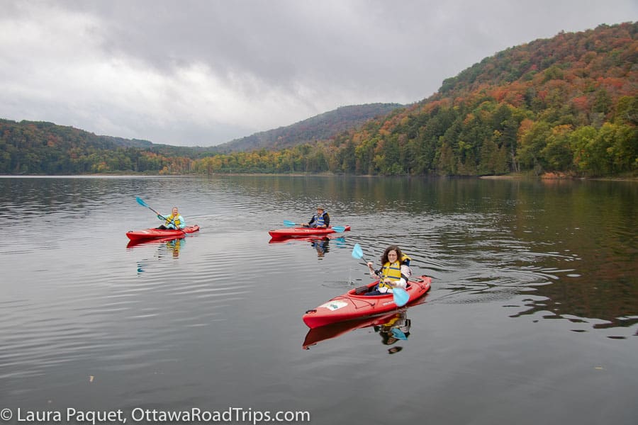 two women and a man in red kayaks with blue paddles on a small lake surrounded by wooded hills with fall colours at kenauk.
