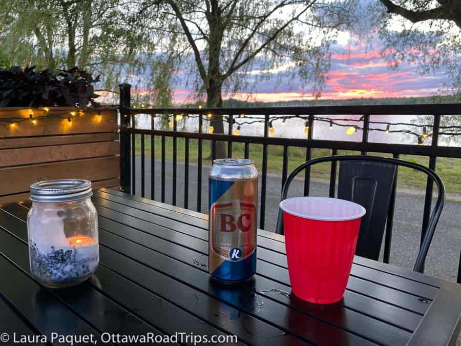 candle, beer can and red plastic cup on metal table, with sunset over river in background