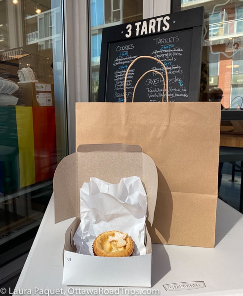 Small pie in a white cardboard box with a brown paper bag and a chalkboard sign in background.