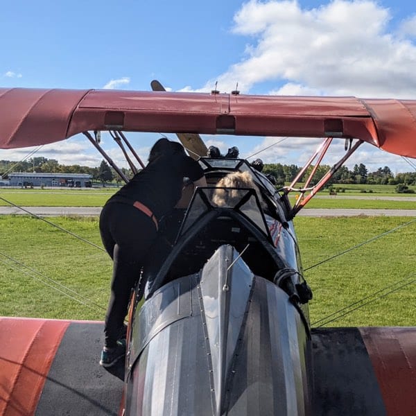 photo from back of biplane showing pilot at left helping woman seated at right fasten seat belt.