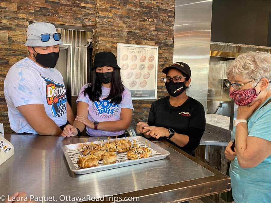 Four people in masks looking at a metal tray of assorted bagels cut into small pieces with toothpicks in them.