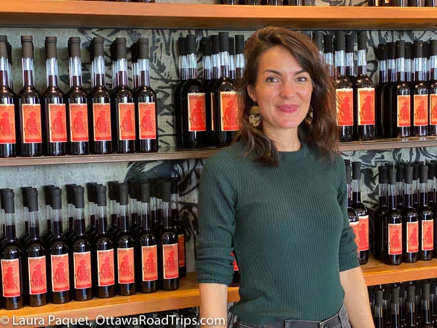 dark-haired woman in green sweater in front of shelves of dark wine with red labels