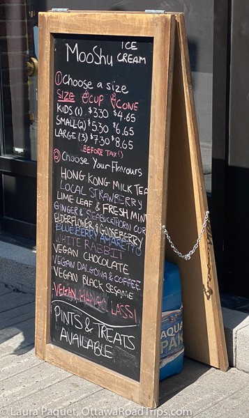 Sandwich board chalkboard listing many flavours of ice cream on sidewalk in front of Moo Shu Ice Cream and Kitchen in Ottawa.