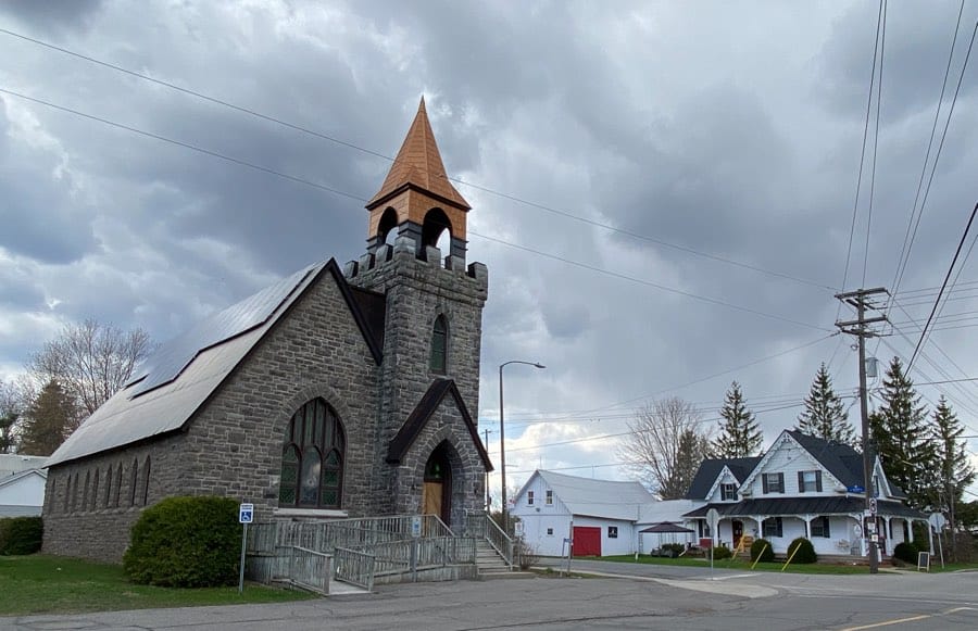 stone church with conical tower and white wooden houses in background at crossroads in navan ontario