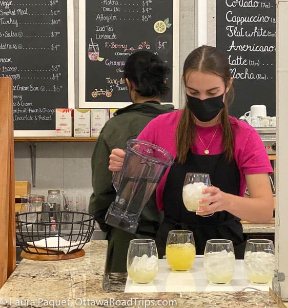 Young woman pouring tea from a blender jug into glasses of ice and lemon syrup, at a granite counter with chalkboard signs in background.