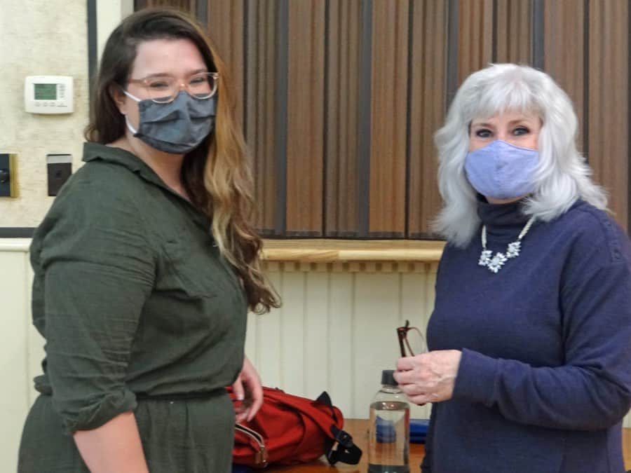 Two women wearing masks in front of a stage with a brown curtain.