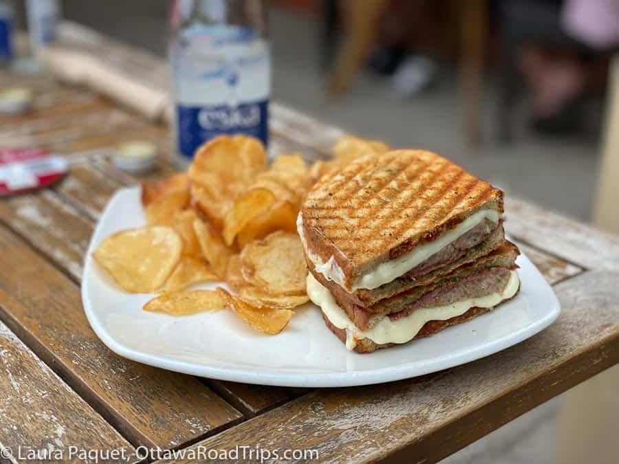 kettle chips and a panini filled with genoa salami and melted cheese, on a white plate sitting on a rustic wooden table.