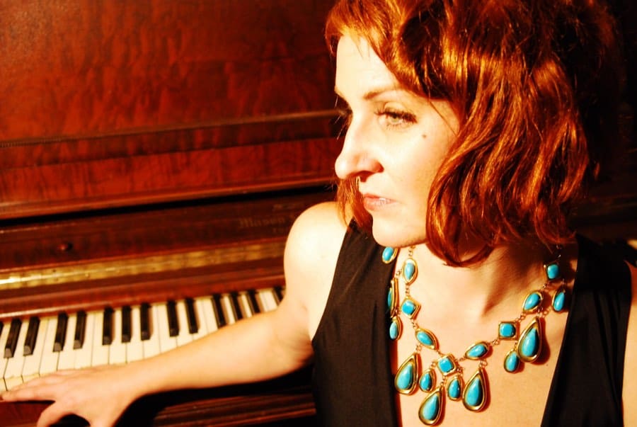 Red-haired woman, wearing a black top and a turquoise necklace, sitting at a piano.