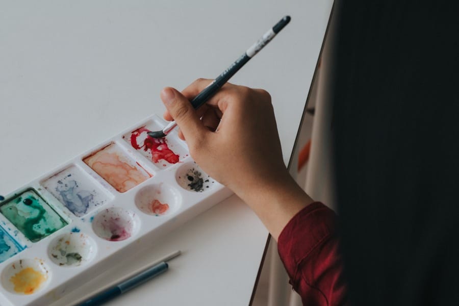 closeup of a hand dipping a paintbrush in a rectangular palette of watercolor paints