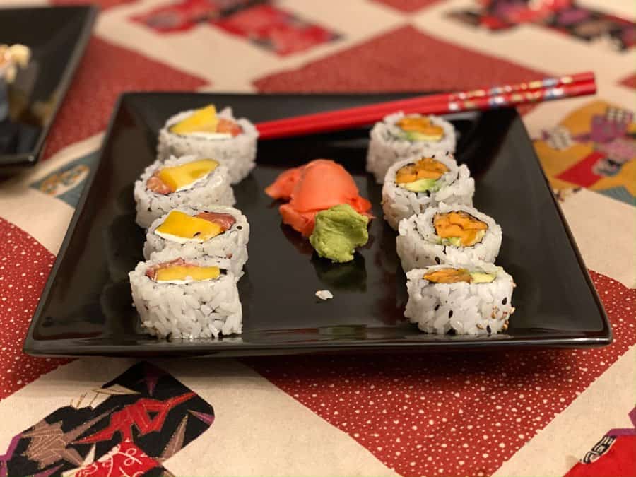 Various sushi rolls and some wasabi on a square black plate on a red tablecloth, with red chopsticks in background.