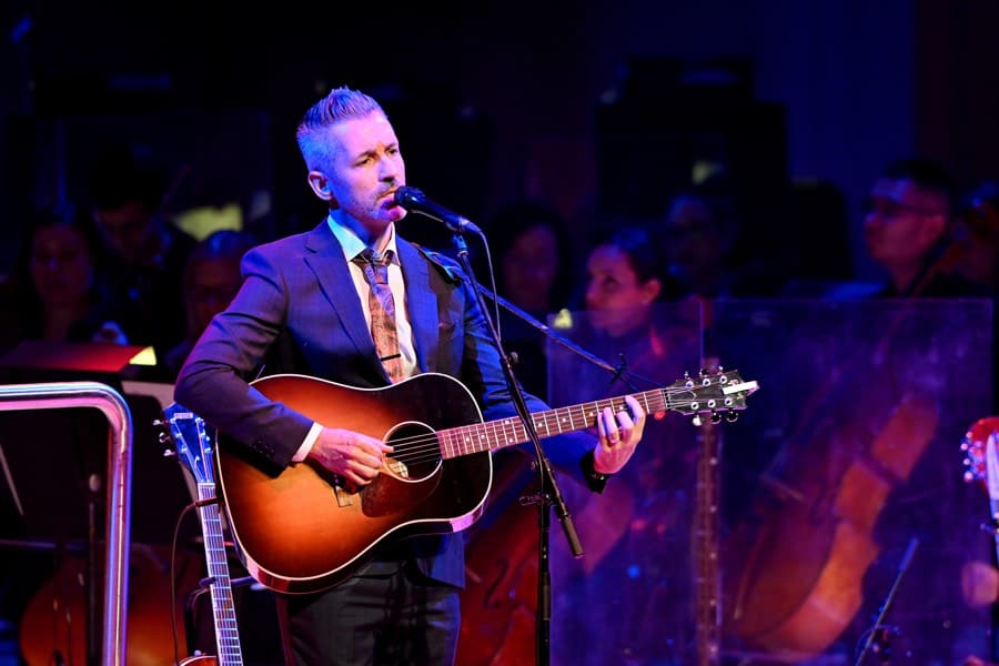 Man in suit and tie playing acoustic guitar with string section of orchestra in background.