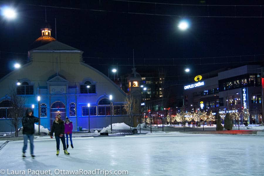 people skating at night on outdoor rink with large yellow building and holiday lights in background